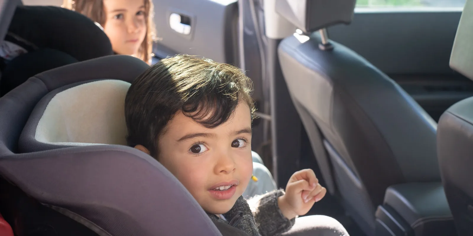 A small boy in a car seat looks round at the camera, an older girl in the seat behind him