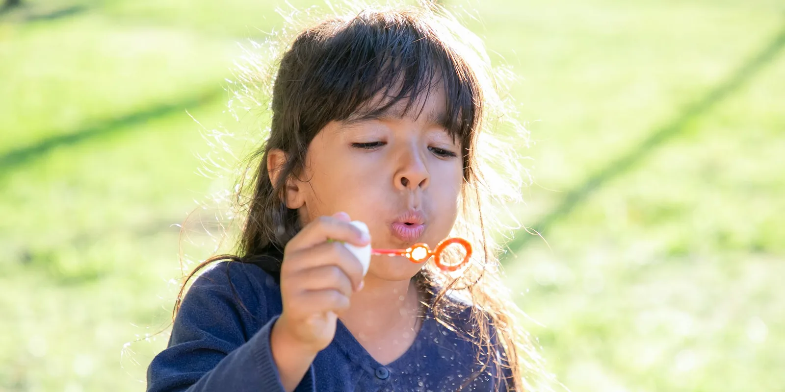 A young girl blows slowly through a bubble wand in a sunny garden