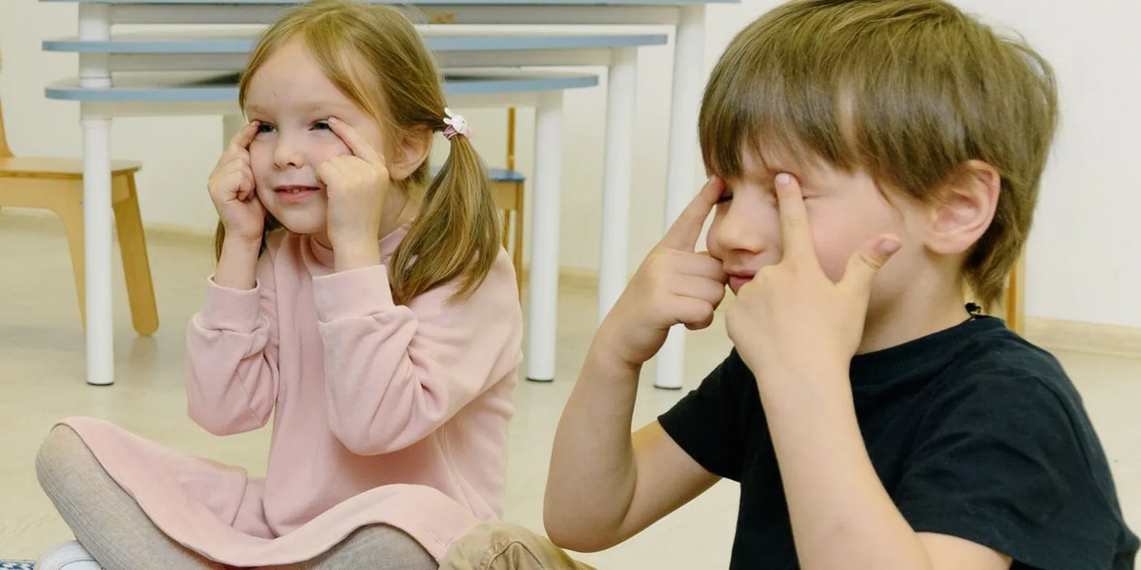 Two young children sitting on a classroom floor press their fingertips to their closed eyes during a calming exercise