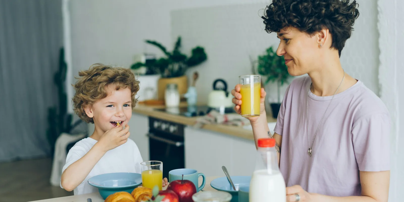 A curly-haired boy laughs over breakfast at the kitchen table while a woman drinks orange juice beside him