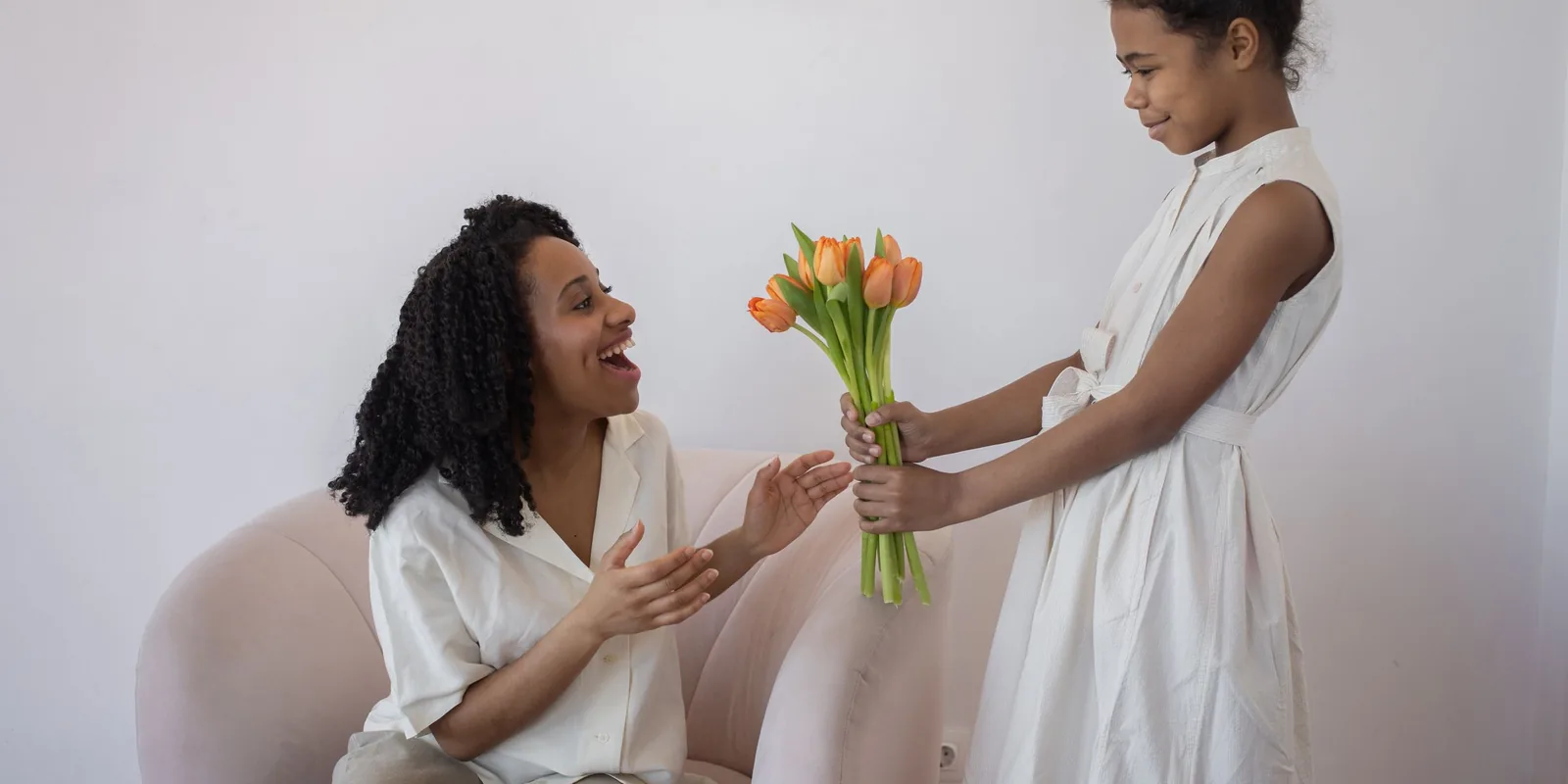 A girl in a white dress hands a bunch of orange tulips to a delighted woman sitting in an armchair