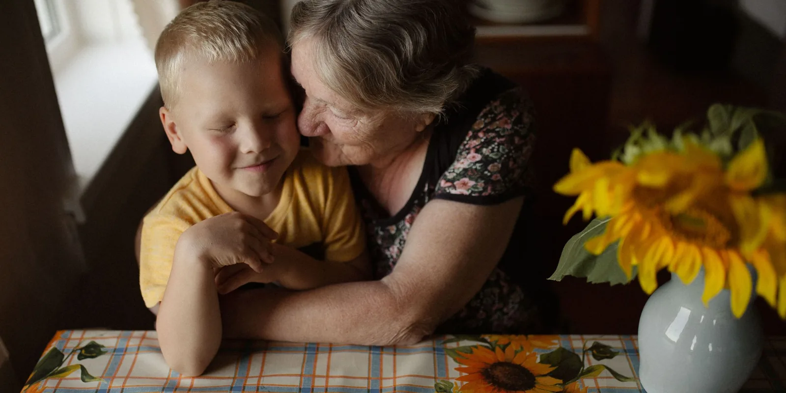 A grandmother cuddles her smiling grandson at a kitchen table beside a vase of sunflowers