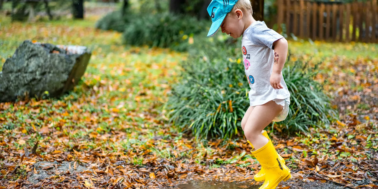 A toddler in yellow wellies stamps in a puddle among autumn leaves