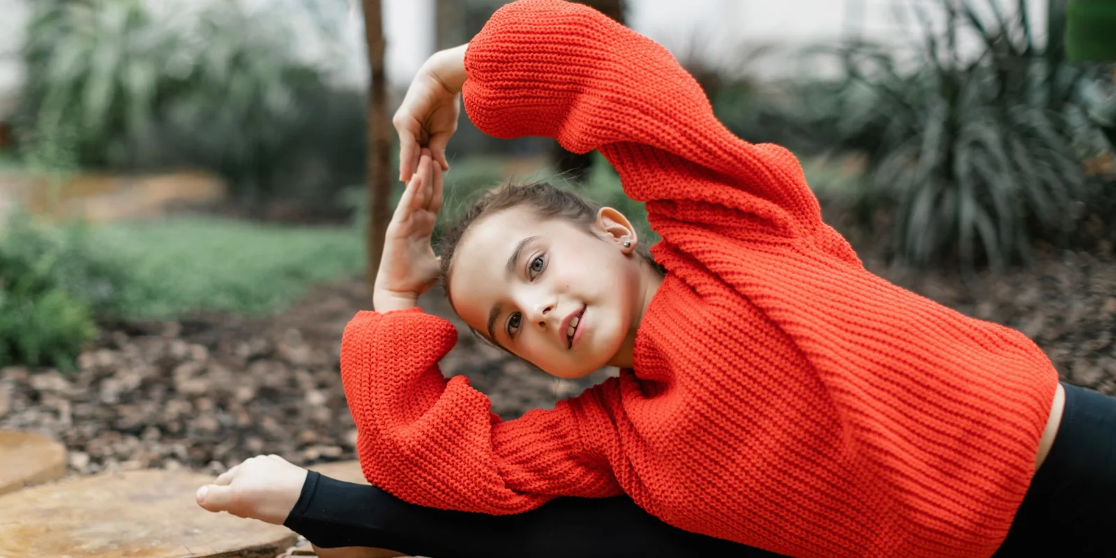 A girl in a red jumper leans into a side stretch with one arm over her head outdoors