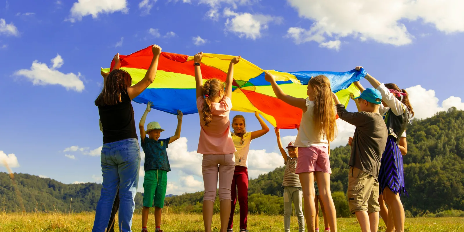 A group of children and adults lift a rainbow play parachute above their heads in a sunny field