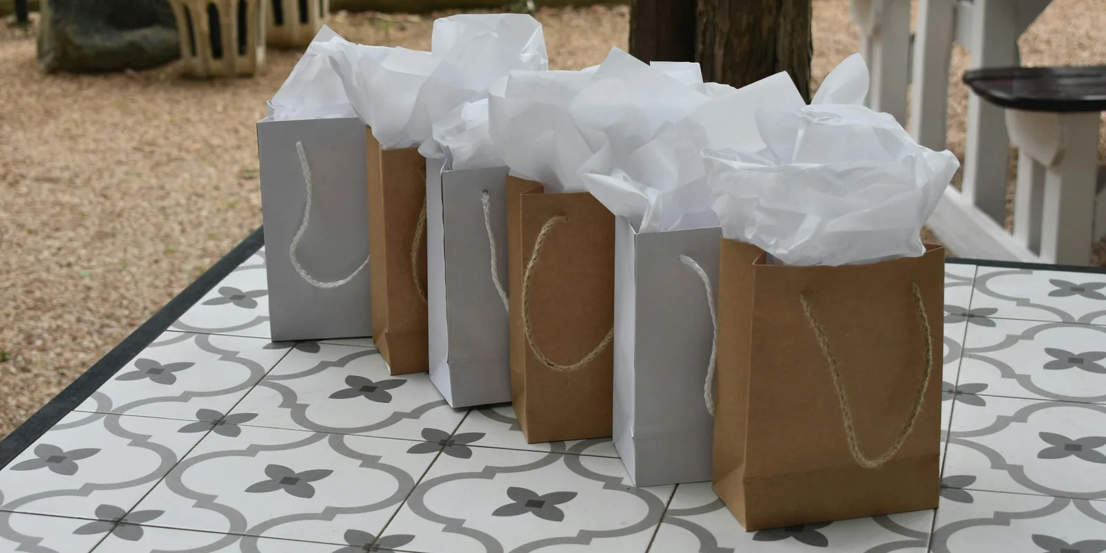 A row of grey and brown paper gift bags stuffed with white tissue paper on a patterned tile table outdoors
