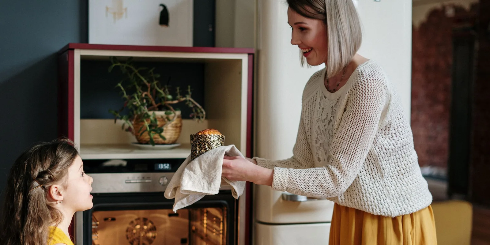 A girl looks up eagerly as a smiling woman lifts a freshly baked cake out of the oven