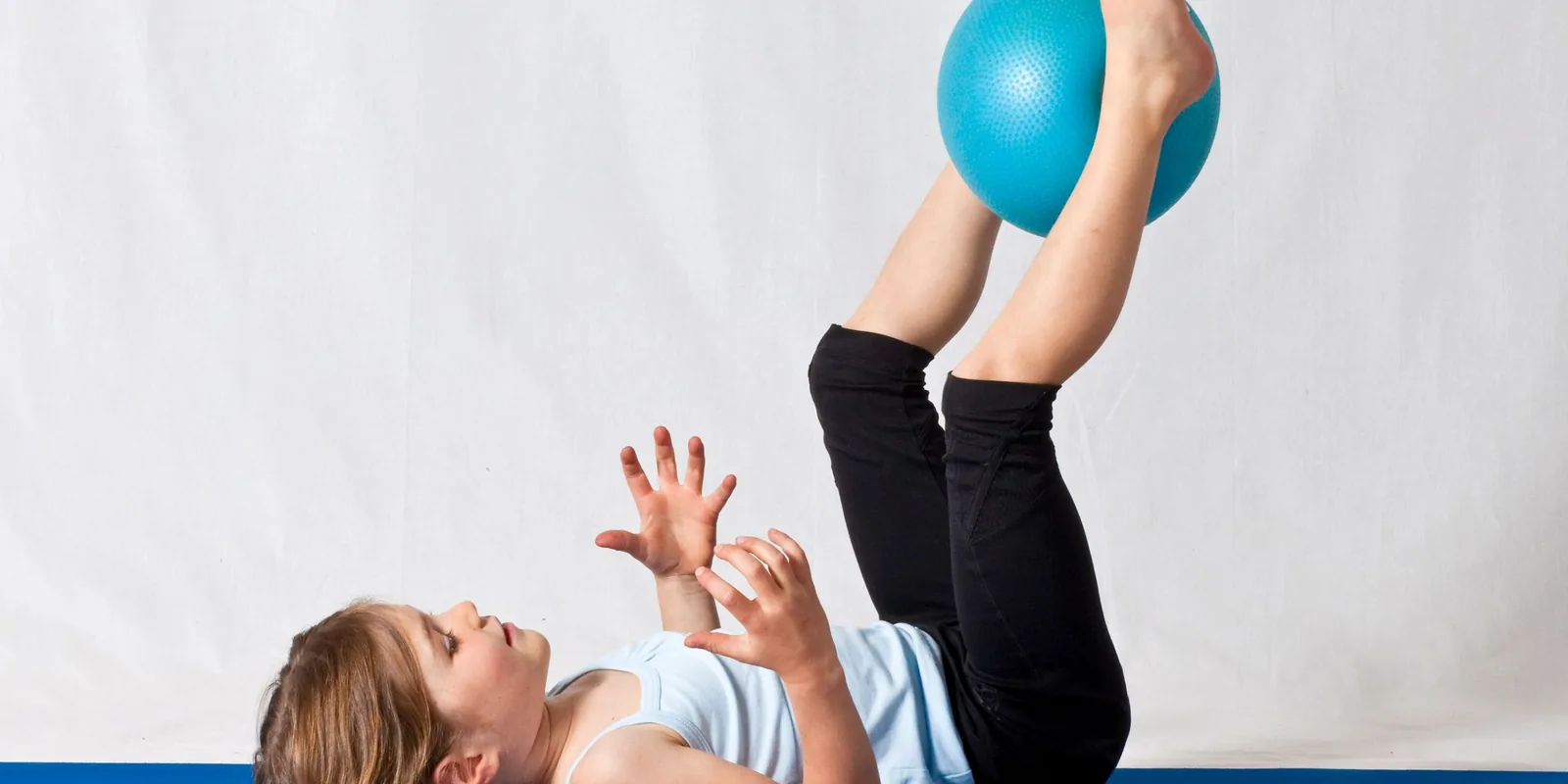 A girl lies on her back and lifts a small blue exercise ball between her feet