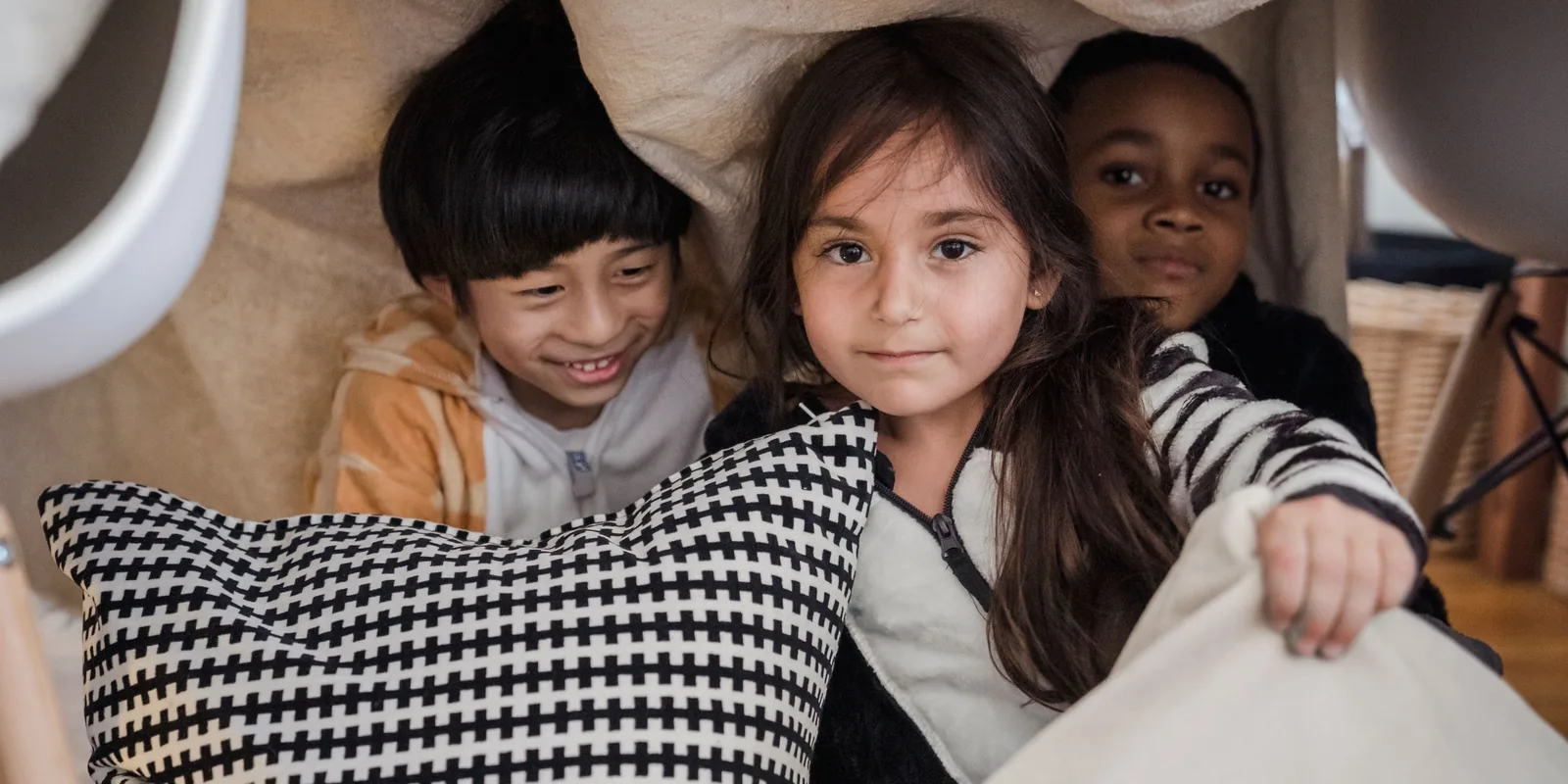 Three children peer out from under a blanket fort draped over chairs, one clutching a cushion