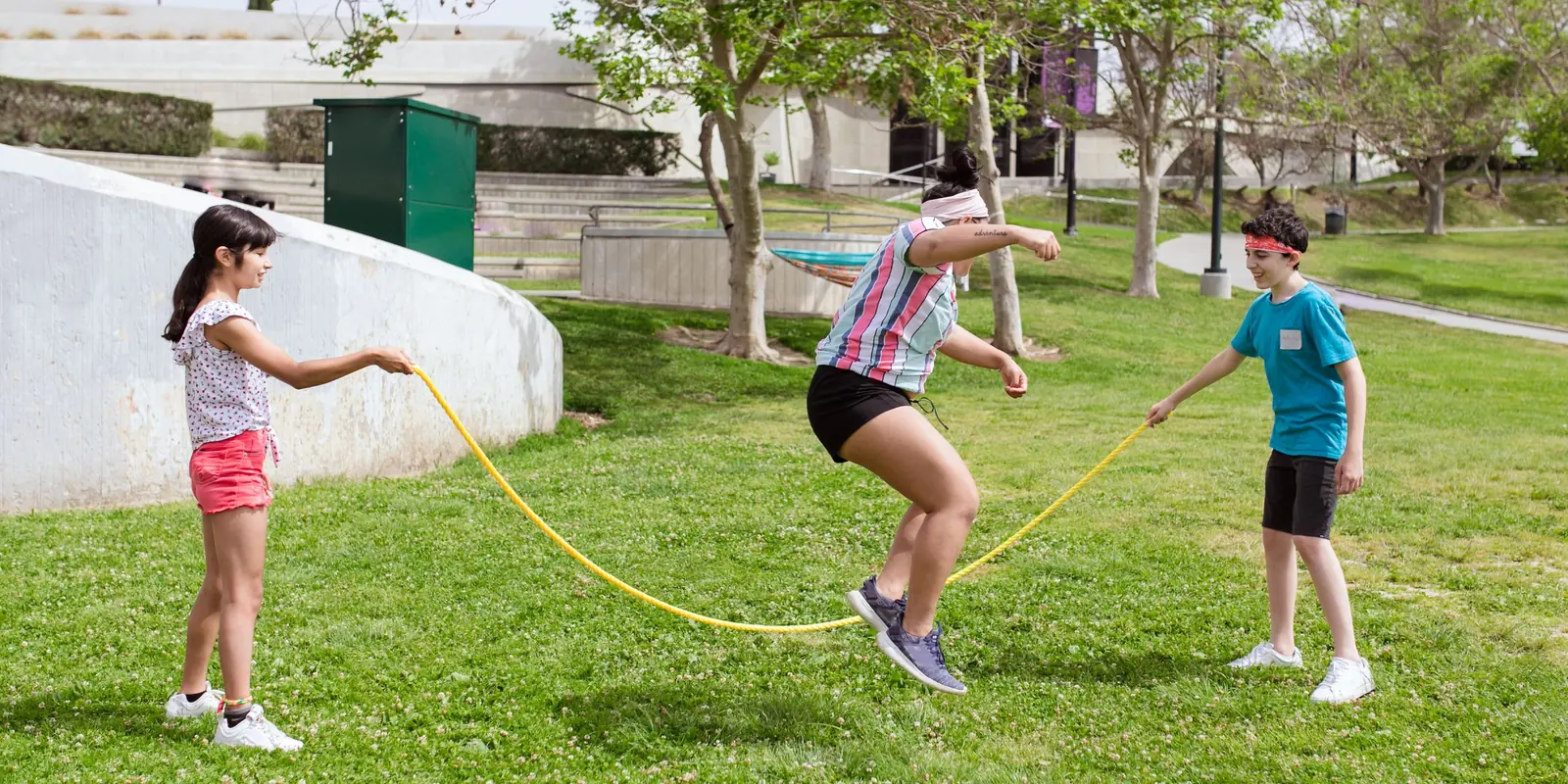 Two children turn a long yellow skipping rope while a third jumps on the grass