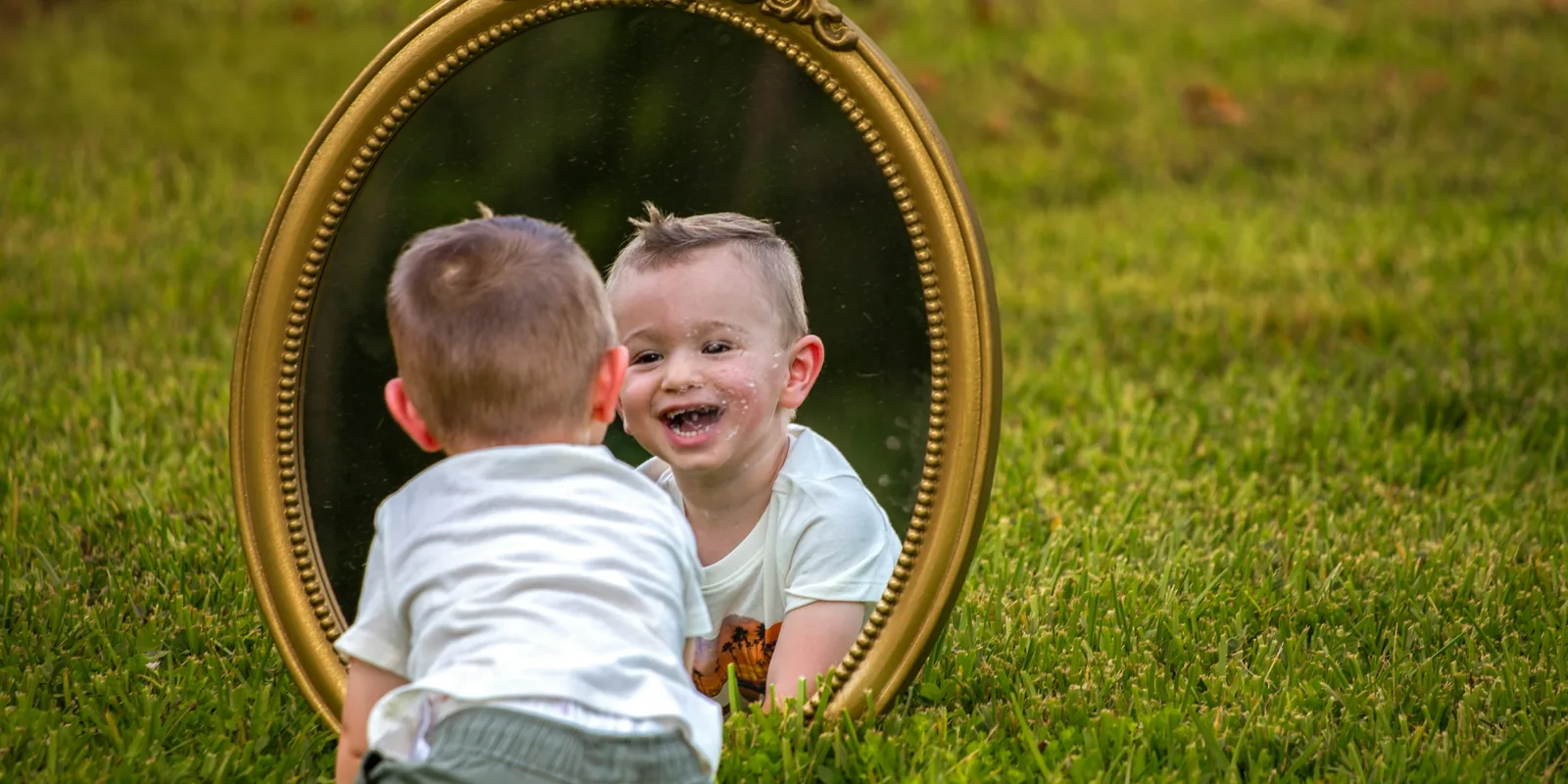 A toddler sitting on the grass grins at his own reflection in a gold-framed mirror