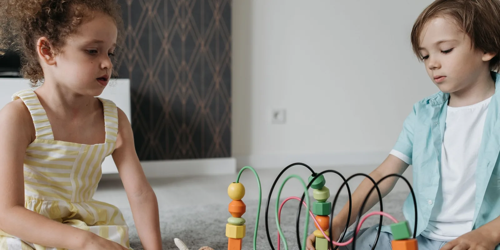 Two young children sit on a living room rug playing with a wooden bead maze