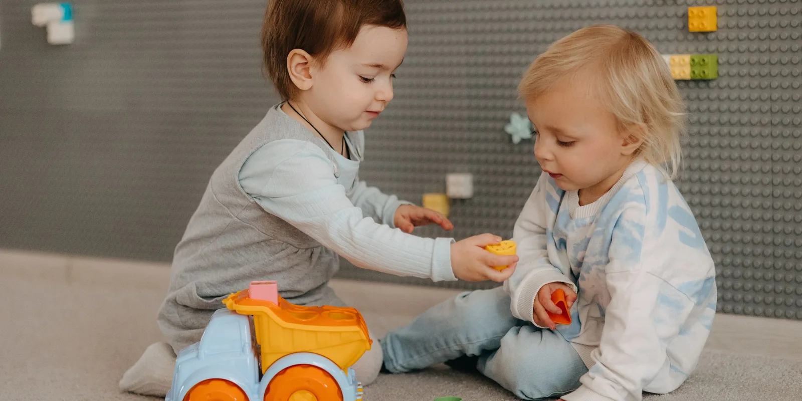 Two toddlers sit on the floor and pass a toy between them beside a toy dump truck