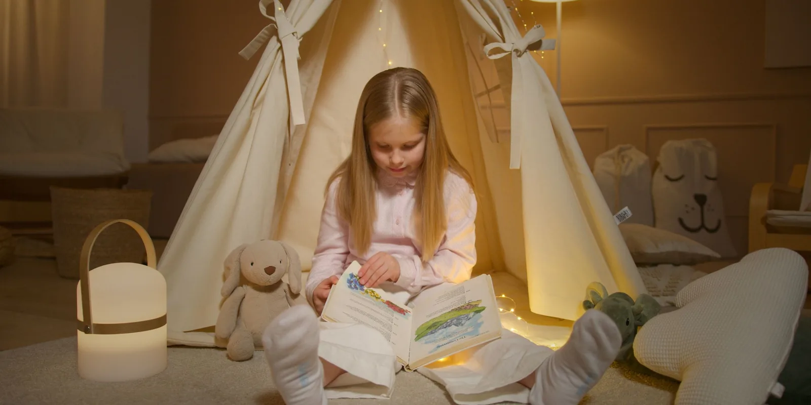 A girl sits at the opening of a canvas teepee reading a picture book by lamplight, a toy rabbit beside her