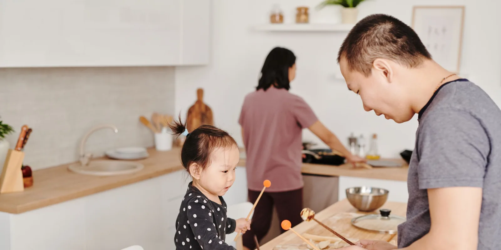 A toddler taps a small drum with orange-tipped beaters at the kitchen counter while two adults cook beside her