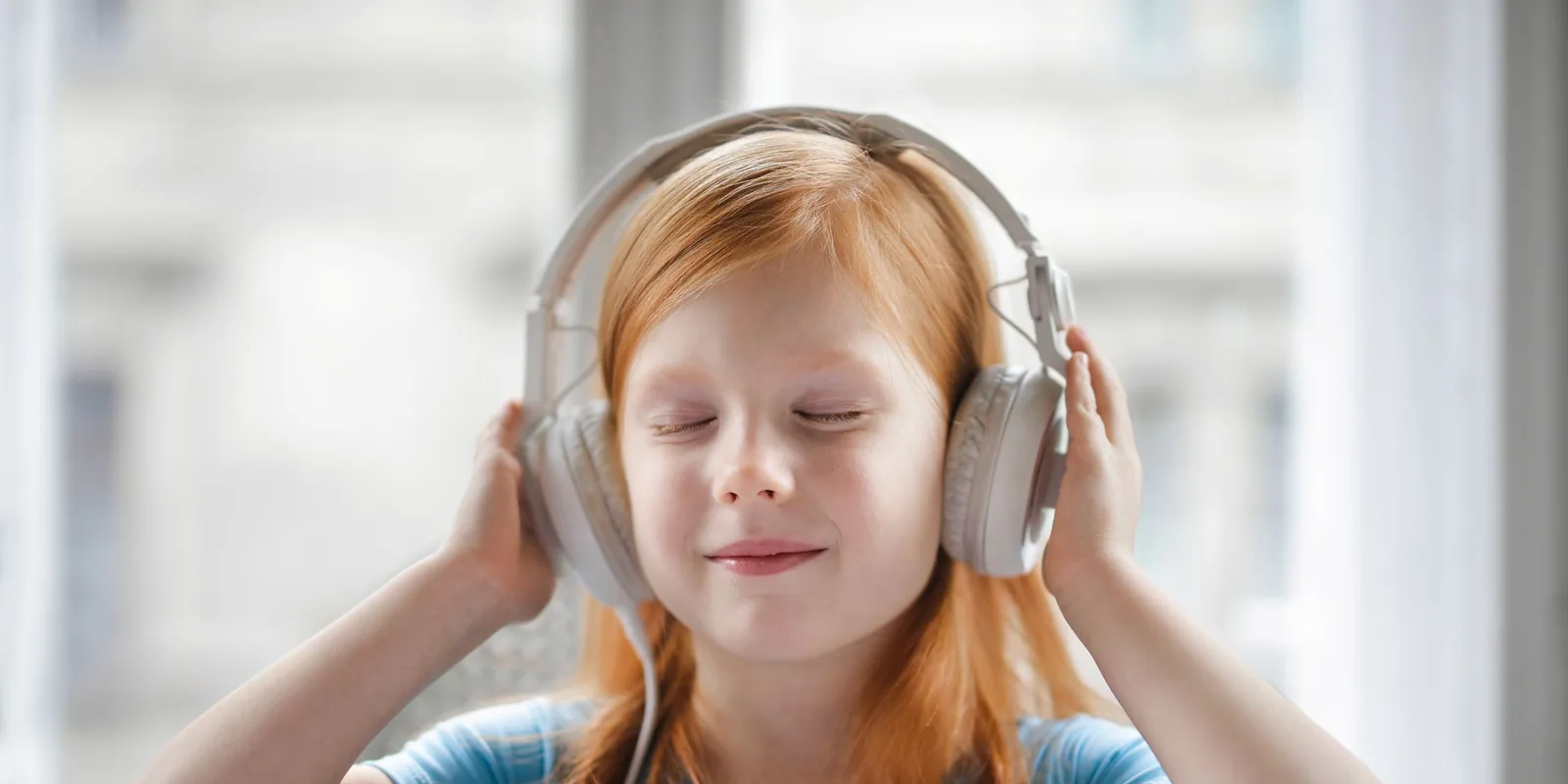 A red-haired girl smiles with her eyes closed, holding a pair of white headphones over her ears