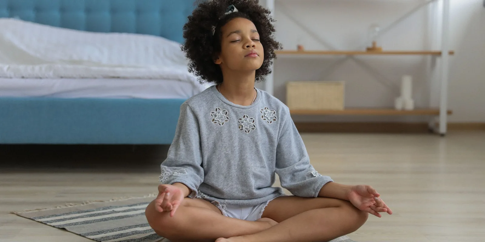 A girl sits cross-legged on a rug in her bedroom, eyes closed and hands resting on her knees