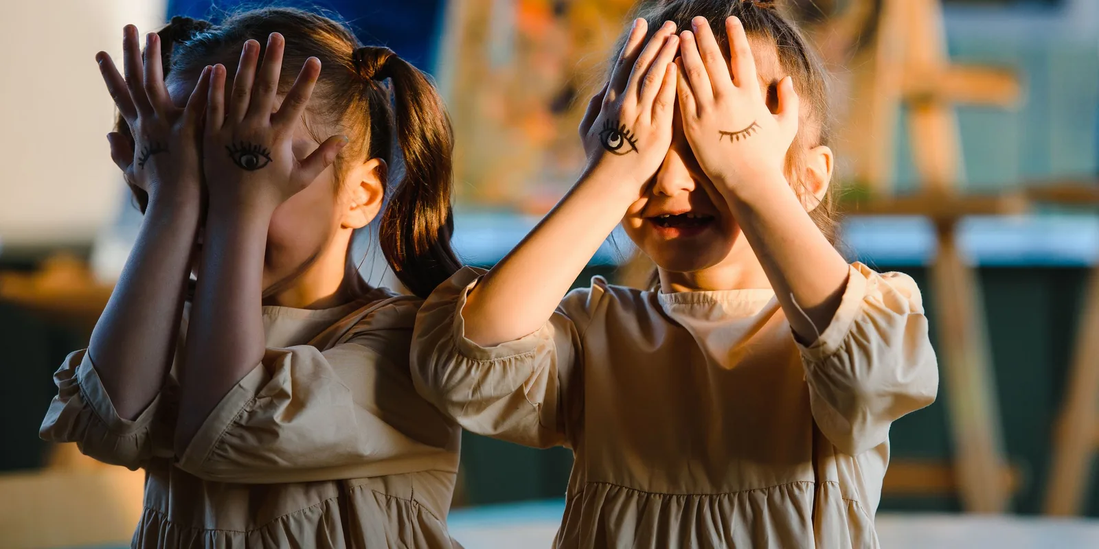 Two small girls cover their eyes with hands that have eyes drawn on them, one laughing