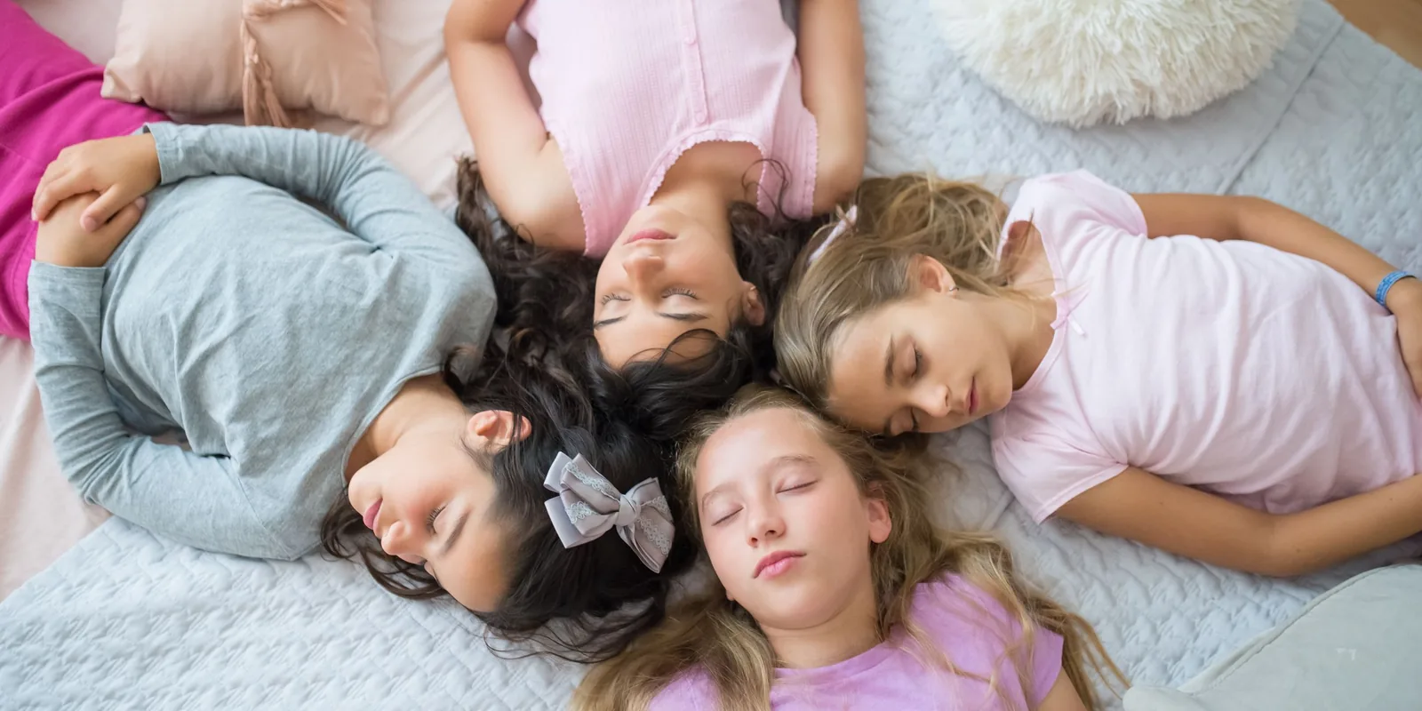 Four girls in pyjamas lie on a bed with their heads together and eyes closed, seen from above
