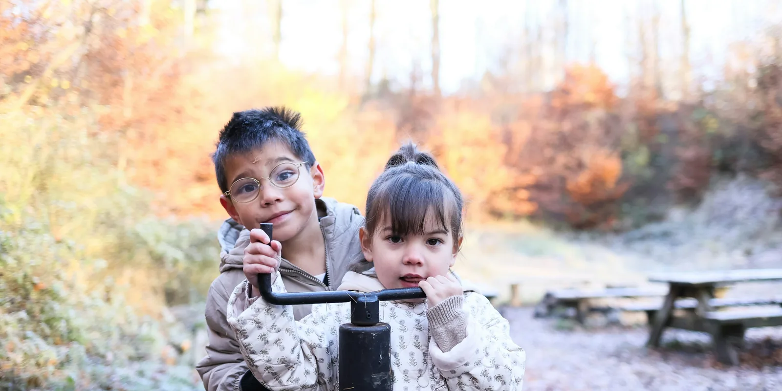 A boy in glasses and a younger girl play at an old water pump in a frosty park