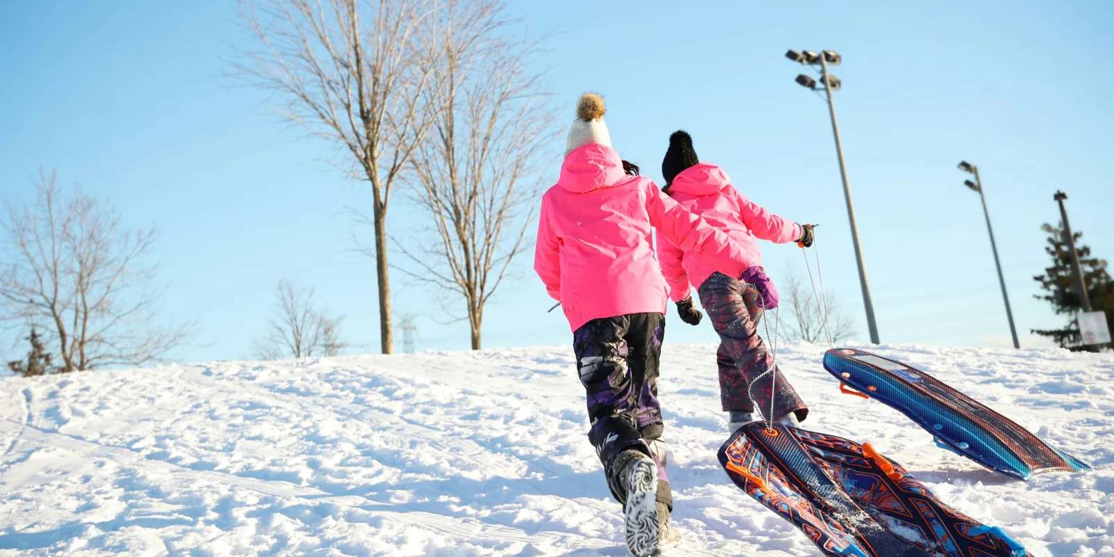Two children in bright pink coats pull their sledges up a snowy hill under a clear blue sky