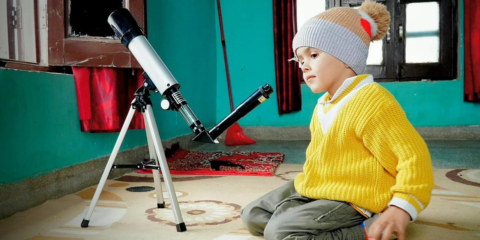 A small boy in a yellow jumper and bobble hat kneels on a rug beside a white telescope on a tripod