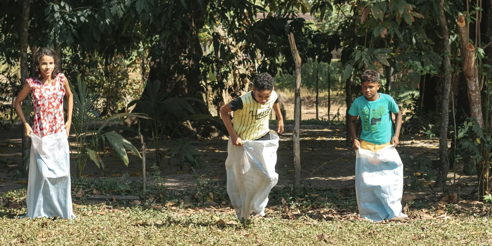 Three children stand in white sacks on a leafy lawn, ready to start a sack race