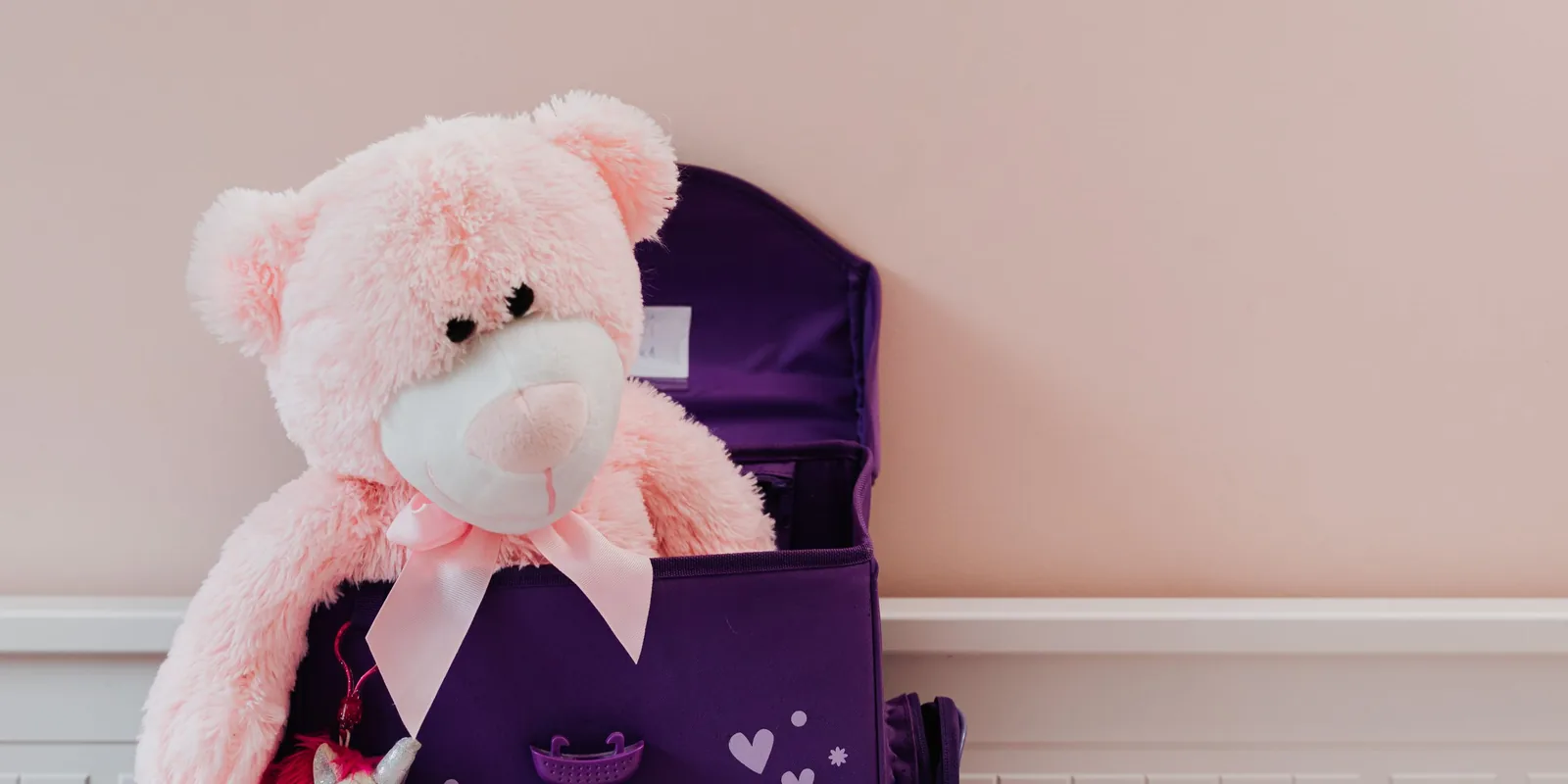 A pink teddy bear with a ribbon bow sits inside an open purple school bag against a pale pink wall