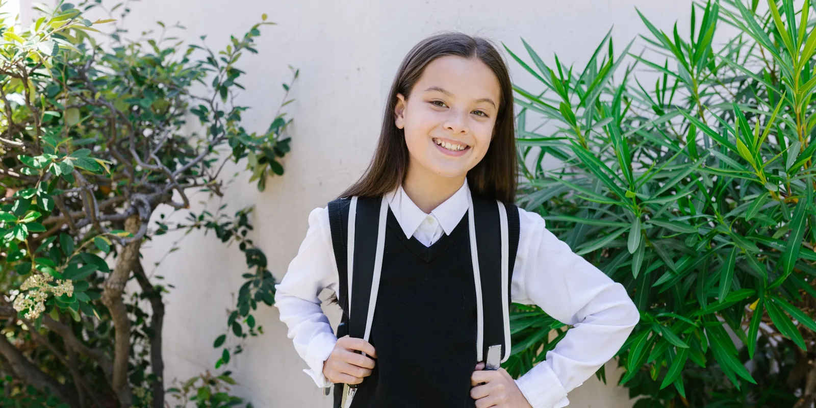 A smiling girl in a white school shirt and black knitted tank top holds the straps of her backpack in front of a garden wall