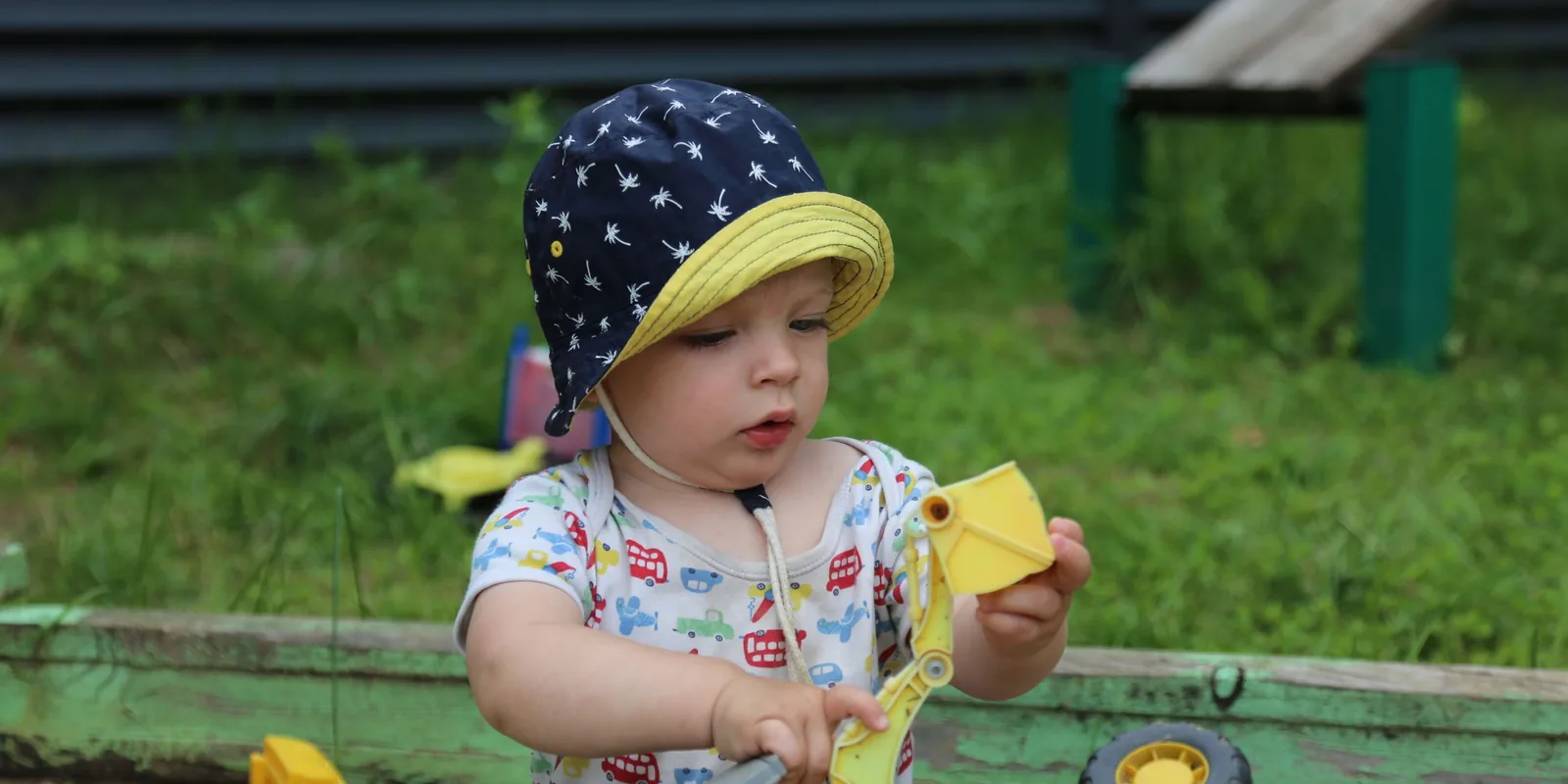 A toddler in a sun hat plays with a yellow digger toy in a sandpit