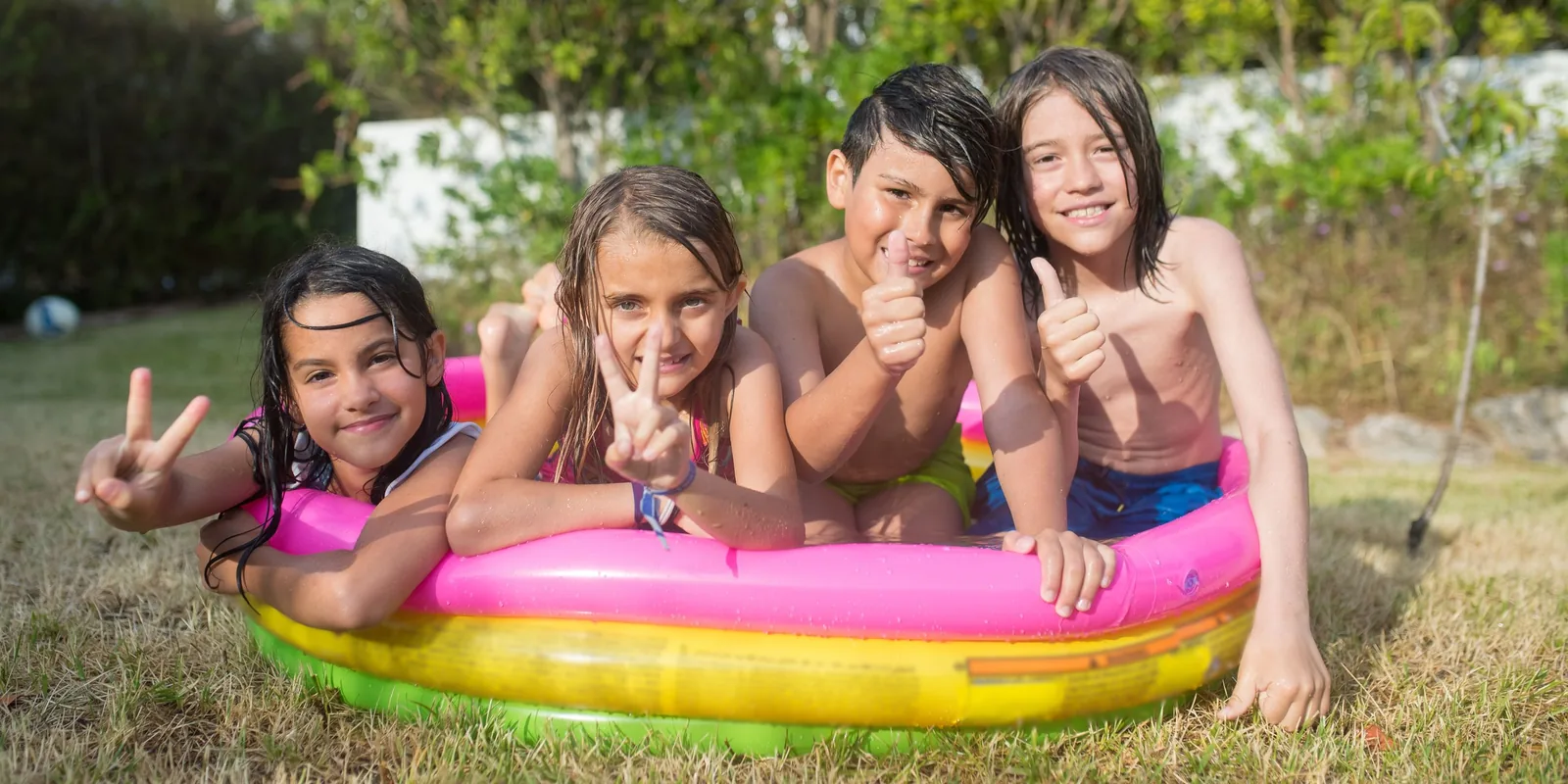 Four children lean over the side of a rainbow paddling pool on a sunny lawn, giving peace signs and thumbs up