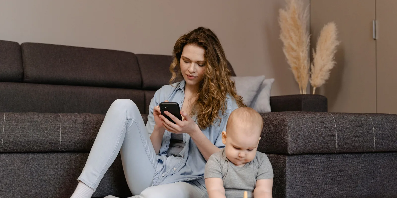 A mother looks at her phone while her baby plays beside her on the floor