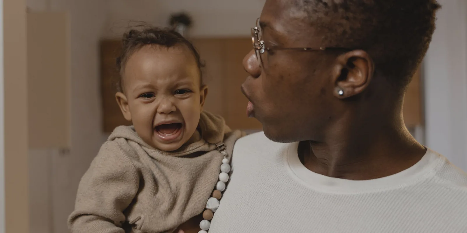 A crying toddler in a hoodie is held by his mother at home