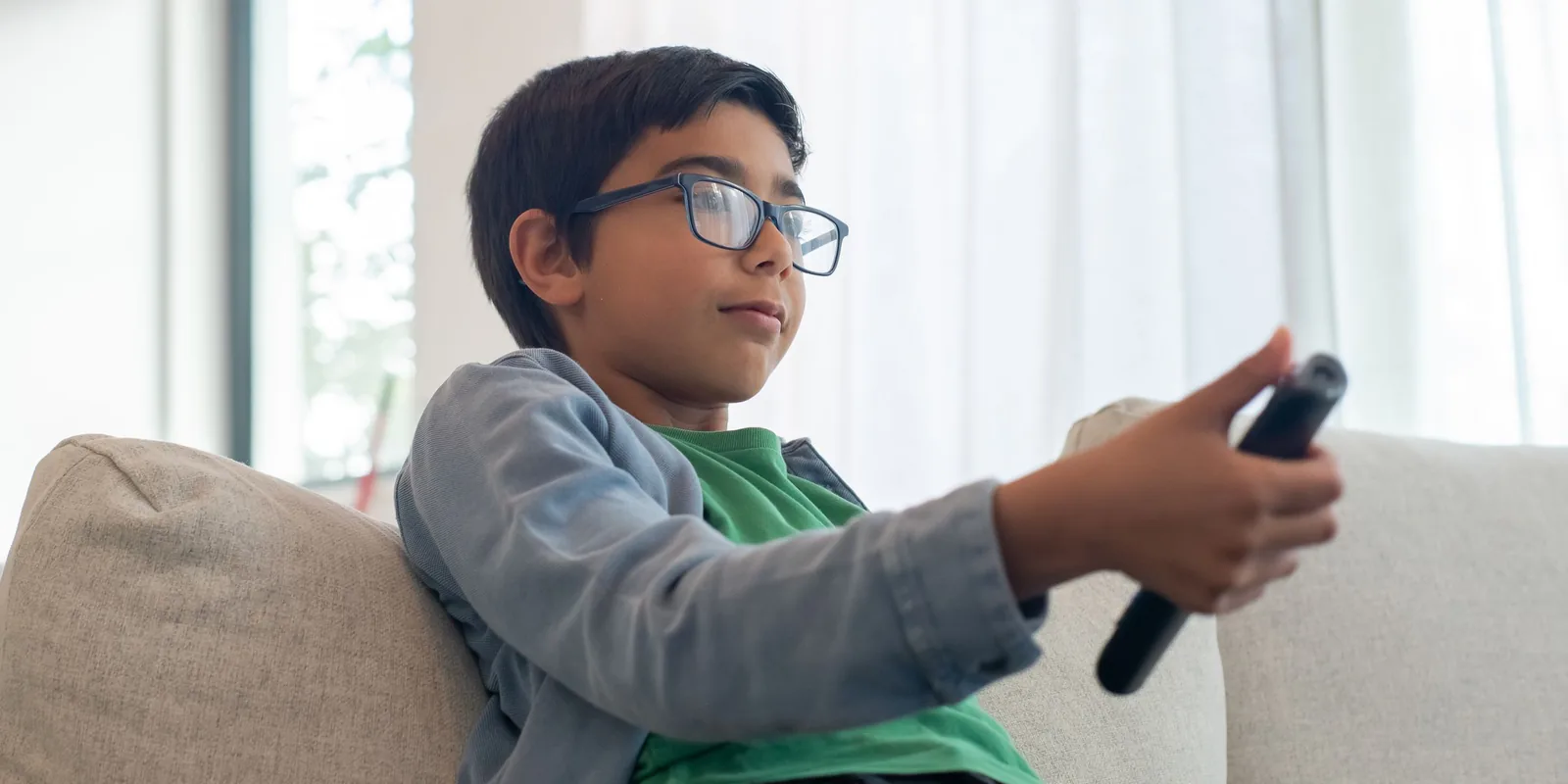 A boy in glasses sits on a pale sofa in a bright room, pointing a TV remote at the screen