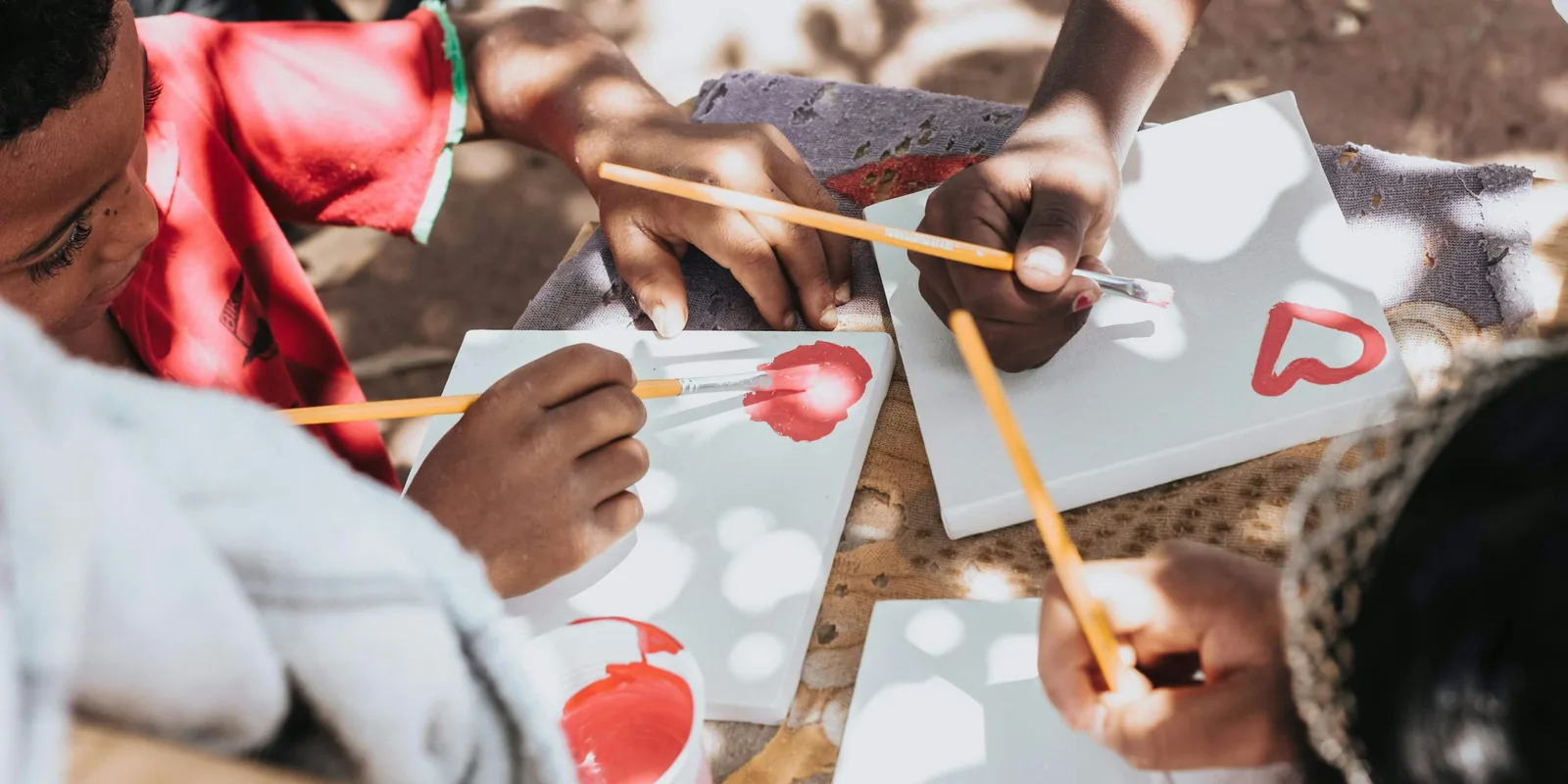 Children paint red hearts on small white canvases with thin brushes, seen from above in dappled sunlight