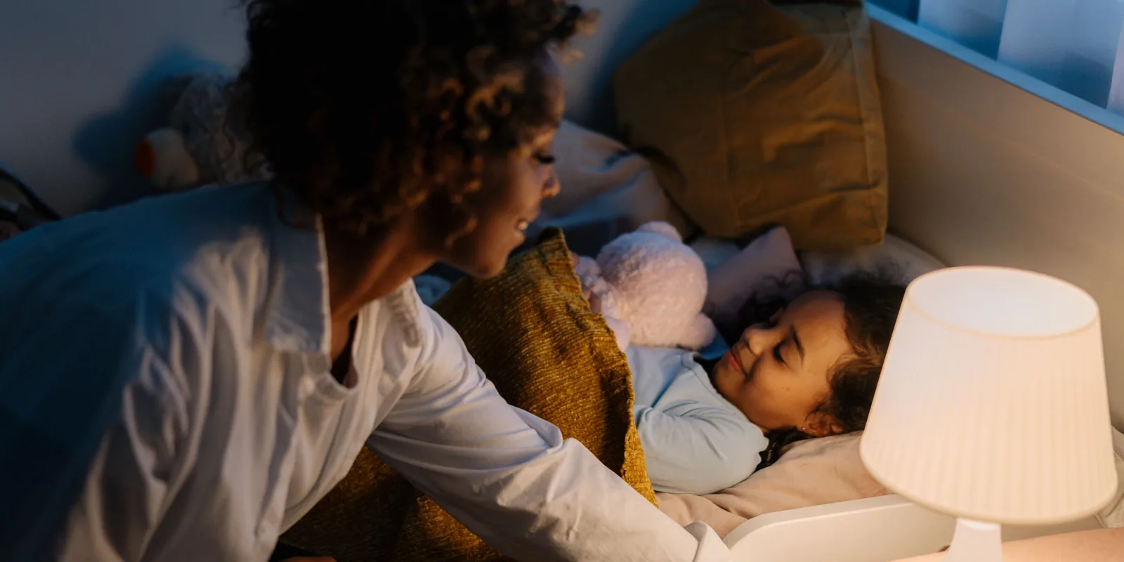 A mother tucks a smiling young girl into bed in the evening, a bedside lamp glowing beside them