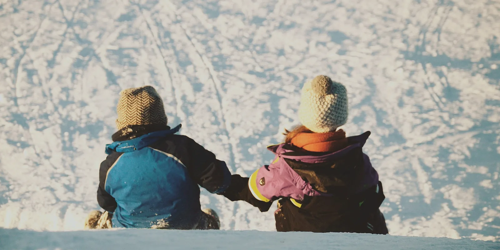 Two small children in snowsuits and woolly hats sit hand in hand at the top of a snowy slope