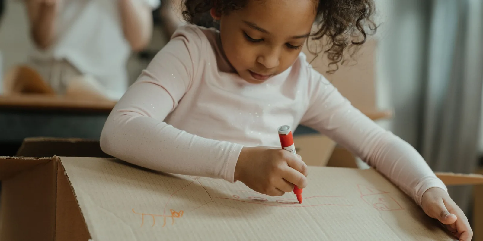 A girl draws with a red pen on the lid of a cardboard box