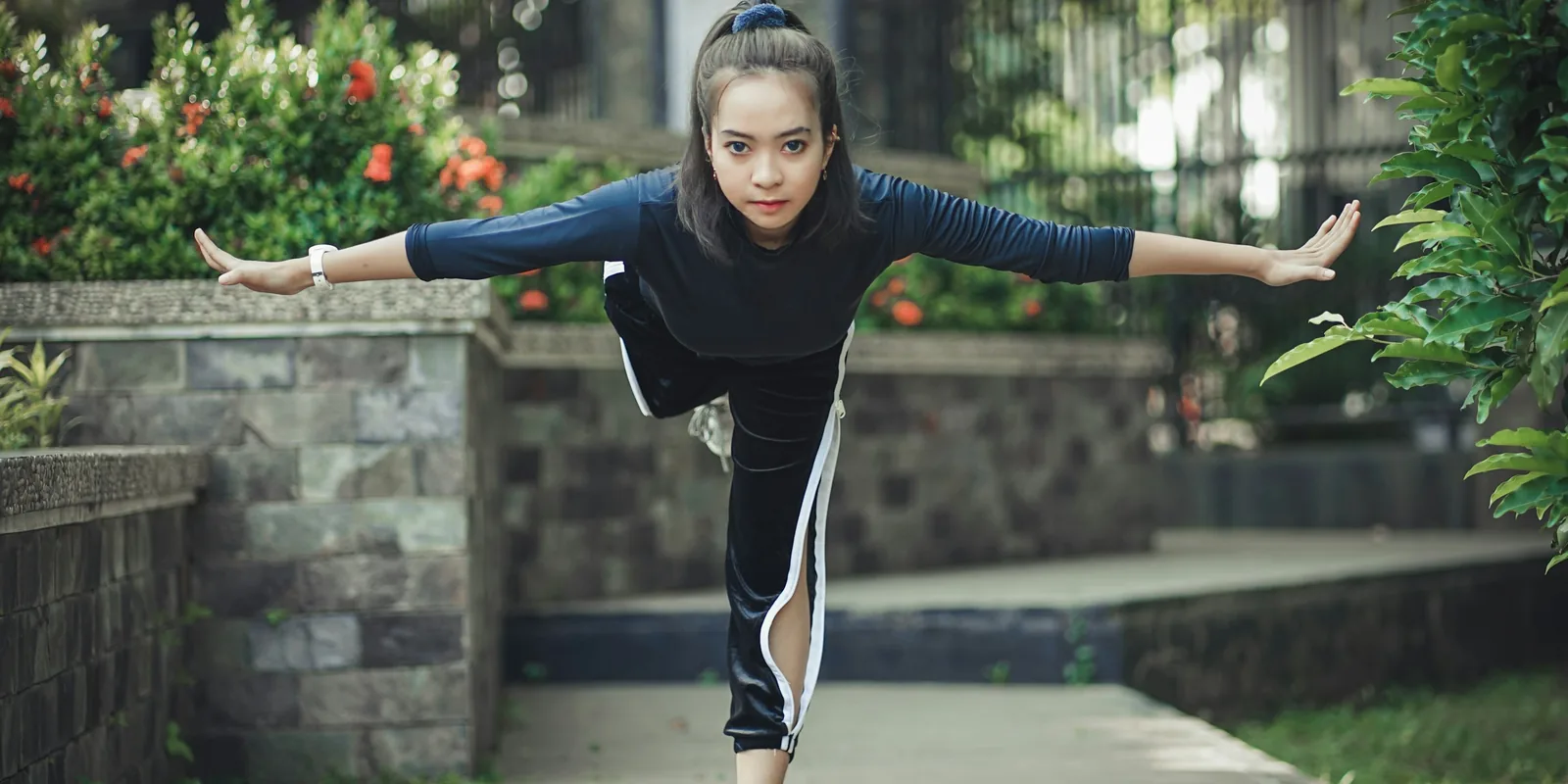 A girl balances on one leg with her arms stretched wide in a garden