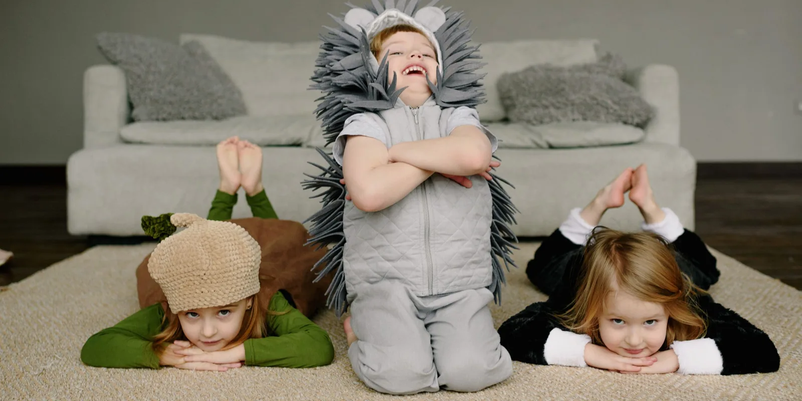 A laughing boy in a hedgehog costume kneels with arms folded between two children in acorn and animal outfits lying on a rug